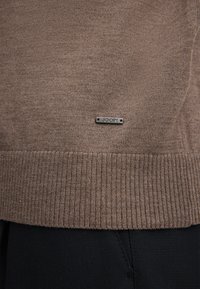 Braun gestrickter Pulloverstoff mit geripptem Saum, versehen mit einem kleinen metallischen Logo-Tag. Schwarze Hose, die am unteren Rand sichtbar ist.