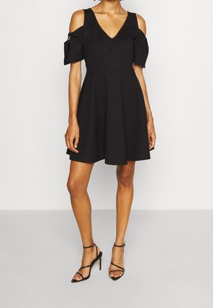 Femme portant une courte robe noire avec des manches épaules dénudées et des sandales noires à talons hauts à brides, debout devant un fond uni.