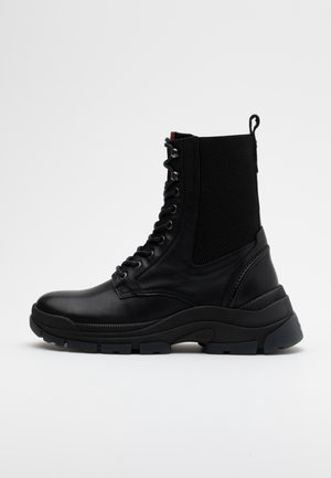 Bottine montante en cuir noir à lacets avec semelle épaisse en caoutchouc et panneau en tissu mesh à l'arrière, vue de profil sur fond blanc.