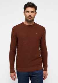 Brauner Strickpullover mit rundem Ausschnitt, gerippten Bündchen und Saum. Verfügt über eine dezente Logo-Applikation auf der Brust. Wird mit blauen Jeans getragen.