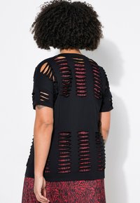 Černé tričko s mnoha svislými výřezy na zádech a rukávech, které odhalují červenou vrstvenou látku pod ním.