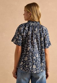 Námornícky modrá kvetinová blúzka s riaseným výstrihom a volánikmi na krátkych rukávoch; obsahuje zložitý biely kvetinový vzor na ľahkej látke.