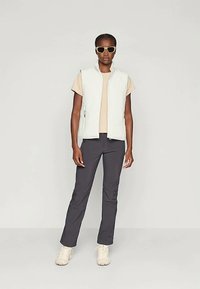 Um colete branco leve e sem mangas sobre uma t-shirt bege, combinado com calças escuras e sapatos de tom claro, acessórios com óculos de sol.