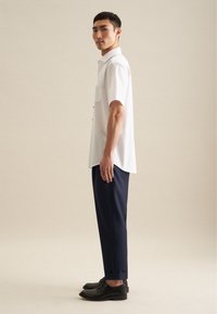 Camicia bianca a maniche corte con taschino sul petto, abbinata a pantaloni slim-fit blu navy e scarpe nere stringate, vista di lato.