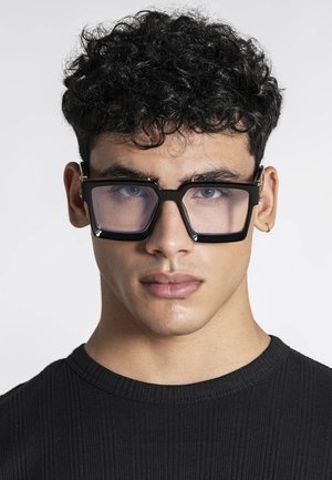 Mladić s kovrčavom tamnom kosom nosi velike crne kvadratne naočale i crnu rebrastu majicu, neutralnog izraza, na svijetloj pozadini.