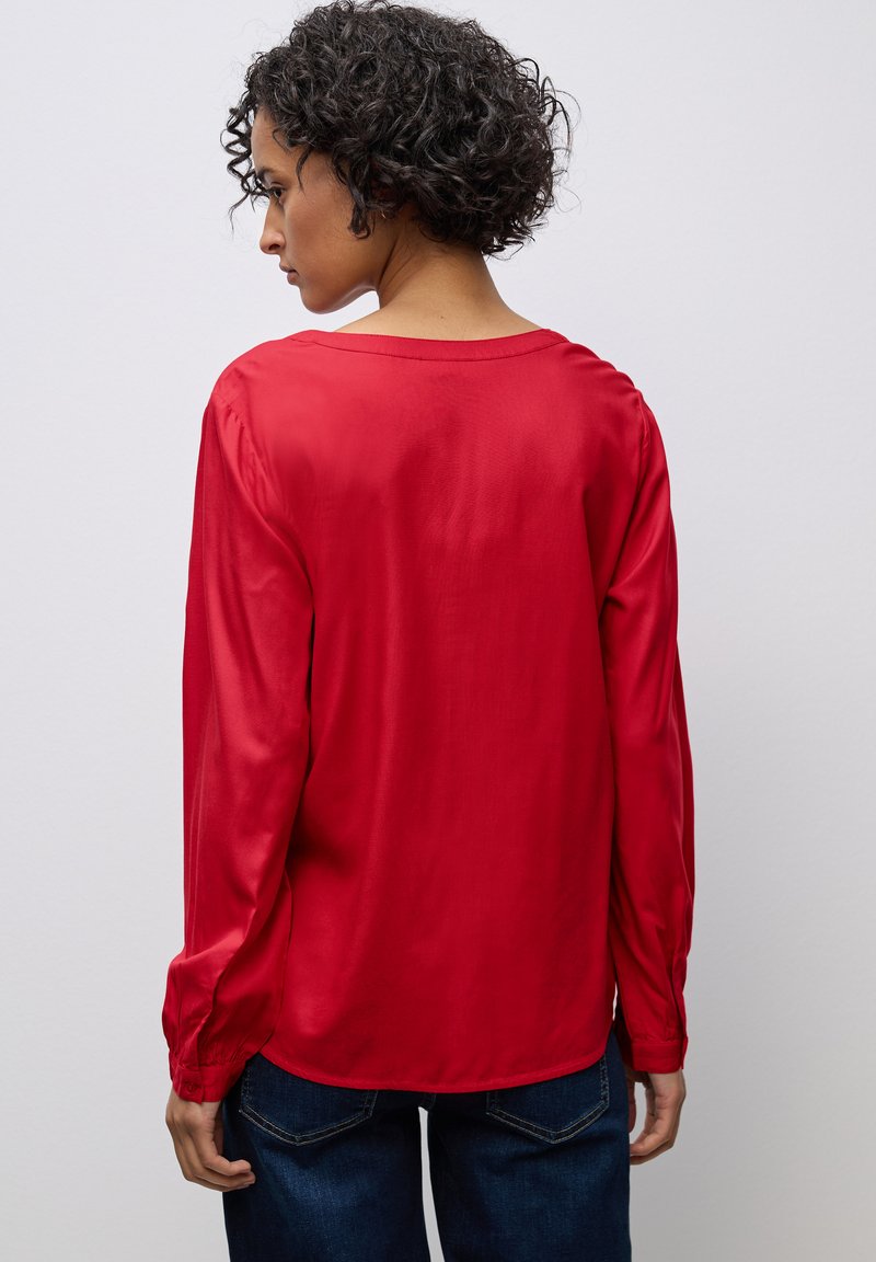 Blusa rossa a maniche lunghe realizzata in tessuto liscio, con scollatura rotonda e polsini arricciati, con un orlo posteriore leggermente più lungo.