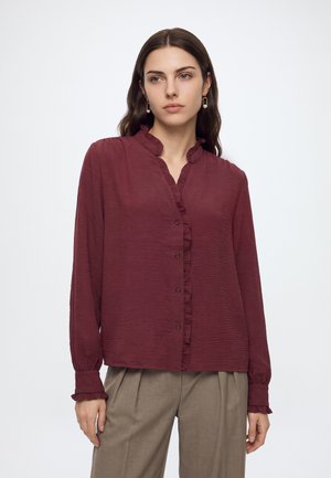 Kvinde med langt brunt hår iført en bordeaux rynket knapbluse og beige bukser, stående mod en ensfarvet hvid baggrund.