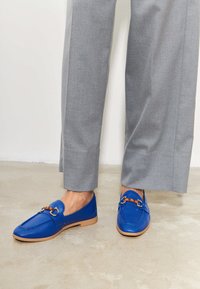 Modré kožené loafers s hnědým akcentem, s dekorativním detailem hardware. Spárované se světlými šedými kalhotami na betonové podlaze.