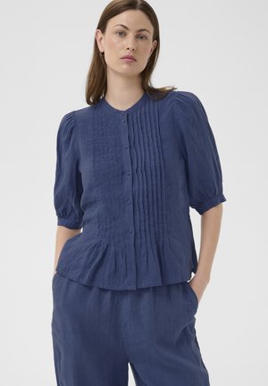 Femme aux cheveux bruns lisses portant un chemisier bleu marine boutonné avec des détails plissés et un pantalon ample assorti, mains dans les poches, fond neutre.