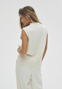 Ärmelloser cremefarbener Pullover aus weichem Material mit geripptem Kragen und Saum. Modell trägt ihn mit hellen Hosen, von hinten gezeigt.