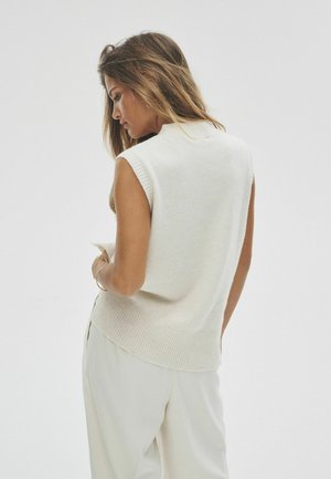 Ärmelloser cremefarbener Pullover aus weichem Material mit geripptem Kragen und Saum. Modell trägt ihn mit hellen Hosen, von hinten gezeigt.