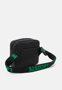 Schwarze Nylon-Crossbody-Tasche mit quadratischer Form, ausgestattet mit einem grünen Logo-Gurt und einer Rücktasche. Reißverschluss oben.