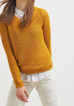 Senfgelber, gestrickter Pullover mit Rundhalsausschnitt und langen Ärmeln, kombiniert mit einem weißen Hemd mit Kragen, das ein schwarzes Rautenmuster aufweist.