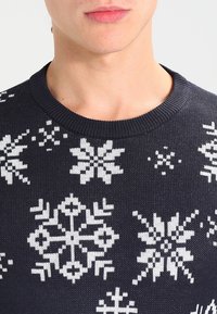 Pull en maille bleu marine avec motifs de flocons de neige blancs. Encolure ronde, tissu texturé et coupe ajustée.