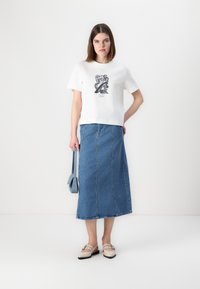 Hvit kortermet t-skjorte med grafisk trykk, kombinert med en blå denim maxiskjørt. Fottøyet er lysebeige med stropper, og en blå veske bæres.