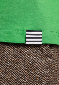 Grøn bomulds t-shirt kant med et sort-hvidt stribet stofmærke, placeret over et struktureret brunt hørstoffer.