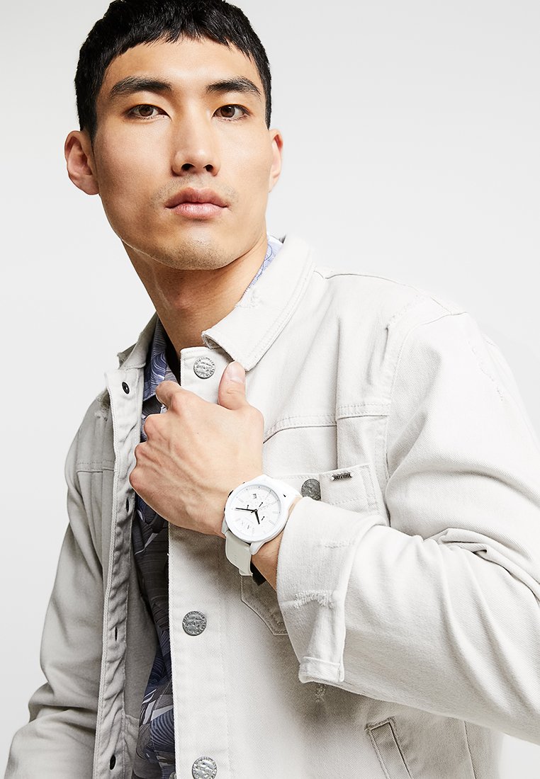 Jeune homme portant une veste en jean claire et une chemise à motifs, montrant une montre-bracelet blanche à son poignet gauche sur un fond uni.