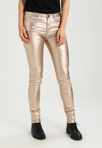 Vaqueros ajustados de tejido dorado metálico con una textura suave, de cintura alta y diseño de cinco bolsillos, combinados con botas negras para crear contraste.