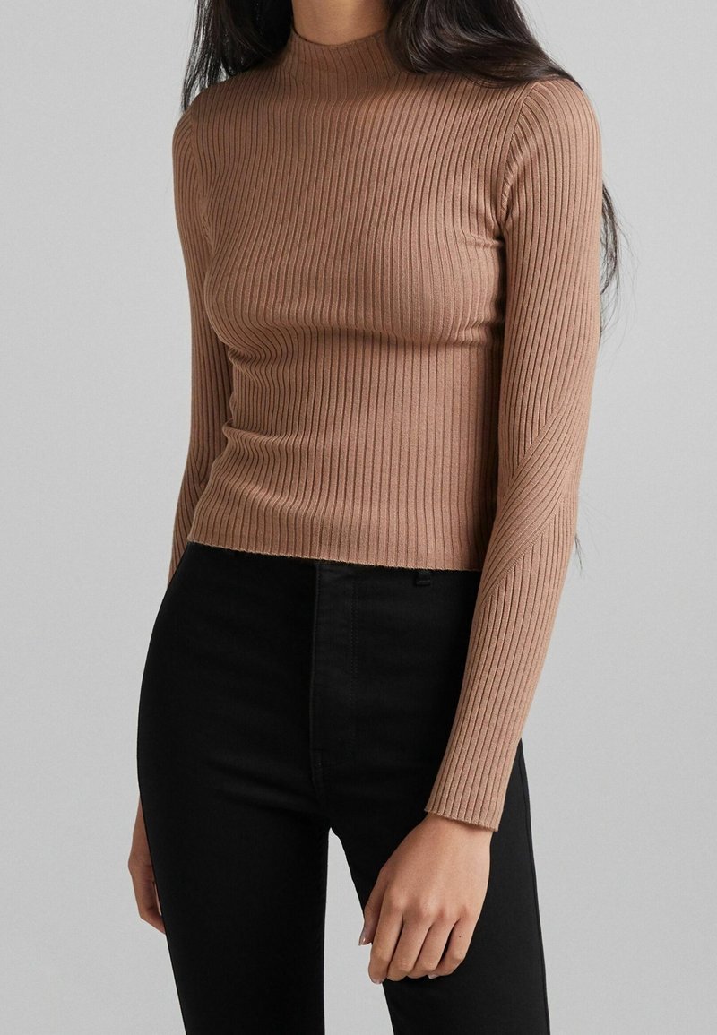 Femme portant un haut beige côtelé ajusté à col roulé à manches longues et un pantalon noir taille haute, sur un fond gris uni.