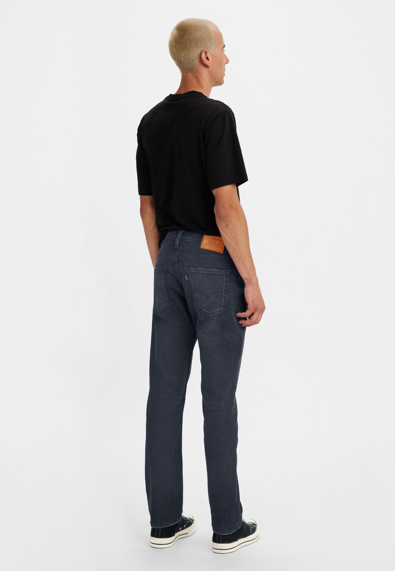 Jean en denim bleu foncé avec une coupe droite, présentant une patch en cuir sur la ceinture arrière, porté avec un t-shirt noir et des baskets.