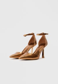 Zlatne metalik štikle s uskim vrhom i remenom oko gležnja. Imaju elegantan dizajn i uski stiletto potplat. Glatka tekstura.