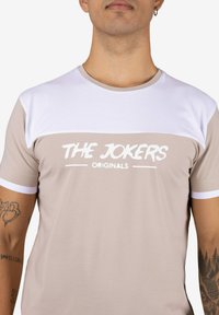 Camiseta beige de manga corta con detalles blancos en los hombros y texto "THE JOKERS ORIGINALS" en blanco en el pecho. Material de algodón suave.