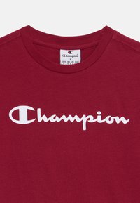 Bourgundy bomullströja med en framträdande vit "Champion"-logotyp. Rund nacke med förstärkt söm. Storleksmärke synligt.