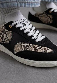 Sorte semskede sneakers med slangeprint-detalje, hvite snører, og en teksturert gummisåle med subtil merkevare på tungen.