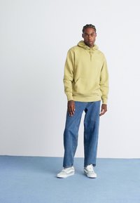 Uomo con capelli intrecciati, indossa una felpa verde, jeans blu e sneakers bianche, si trova su un pavimento blu contro un muro bianco semplice.