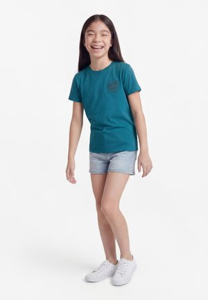Lachend meisje met lang donker haar, gekleed in een turquoise T-shirt met korte mouwen, lichte spijkershorts en witte sneakers, staand voor een witte achtergrond.