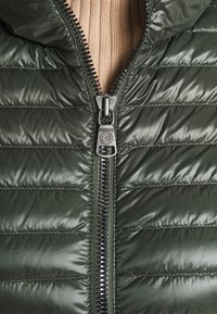 Detailní záběr na částečně rozepnutou tmavě zelenou prošívanou bundu přes béžový žebrovaný svetr, ukazující kovový zip a texturu polstrované látky.