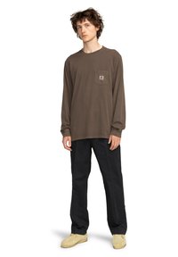 Hnedé tričko s dlhým rukávom a vreckom; kombinované s tmavými nohavicami a svetlými topánkami. Hladká tkanina, voľný strih s detailom loga.