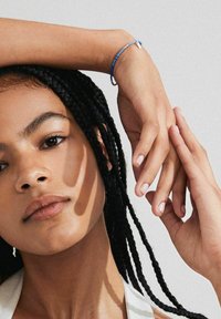 Brazalete de cuentas azules en la muñeca, manos finas con uñas naturales, cabello trenzado y rasgos faciales suaves sobre un fondo neutro.