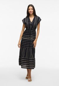 Smiling woman in a cap-sleeve, black patterned maxi dress stands against a light background, showcasing flow and design with beige sandals.