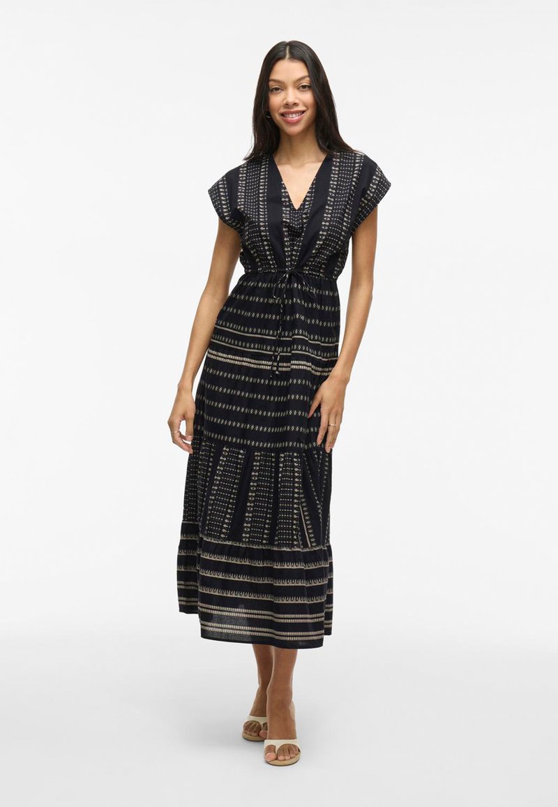 Smiling woman in a cap-sleeve, black patterned maxi dress stands against a light background, showcasing flow and design with beige sandals.