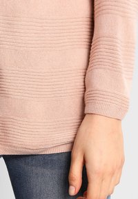 Maglione rosa lavorato a maglia con texture a coste orizzontali e orlo arrotondato. Il primo piano mostra parte di una mano e jeans di denim.