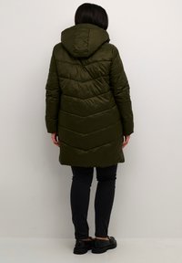 Olijfgroene pufferjas met een capuchon, voorzien van een chevronpatroon en een middellange lengte, gecombineerd met donkere jeans en schoenen.