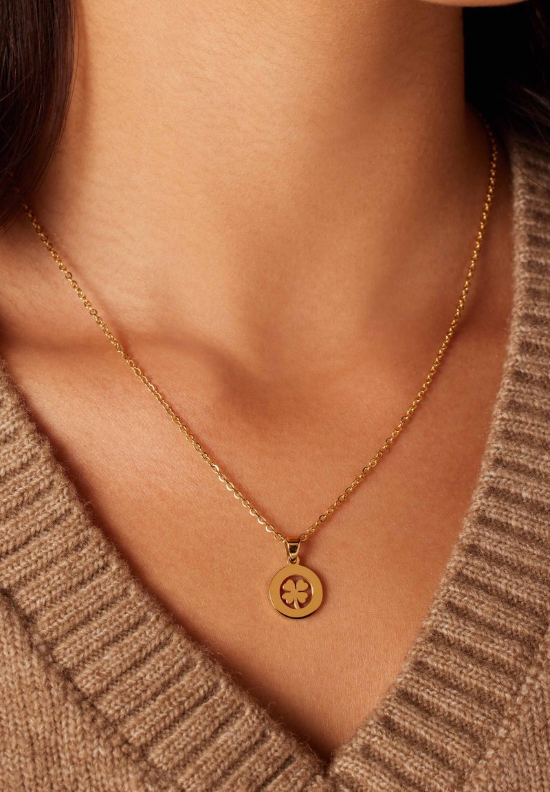 Gouden ketting met een ronde hanger met een klaverthema, bevestigd aan een fijne ketting, gedragen over een beige gebreide trui.
