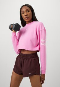 Rózsaszín, cropped kapucnis pulóver, fehér Nike logóval az ujjon, barna, elasztikus derékbőségű rövidnadrággal párosítva, amelyen egy kis szürke Nike swoosh található. Fekete hatszögletű kézi súlyzók.
