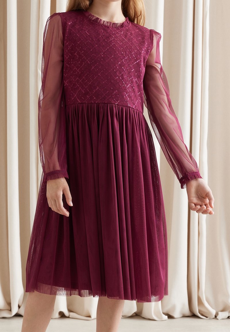 Fille portant une robe bordeaux longueur genoux avec des manches longues transparentes et un corsage texturé, debout devant des rideaux crème.