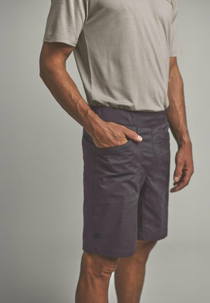 Man wearing beige short-sleeve shirt and dark knee-length shorts with hand in front pocket against neutral background.