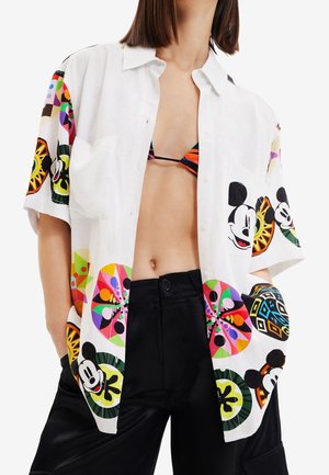 Kobieta ubrana w białą, rozpiętą koszulę z krótkim rękawem, zdobioną kolorowymi okrągłymi wzorami i twarzami Myszki Miki, w połączeniu z czarnymi spodniami i wielokolorowym bikini.