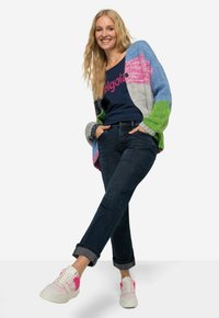 Cardigan a maglia multicolore con sezioni blu, rosa, verde e grigio. T-shirt blu navy con testo rosa, jeans in denim scuri e sneaker bianche con accenti rosa.