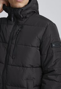 Black puffer jacket with a high collar. Features a front zipper, a chest pocket, and a textured fabric with stitched panels.