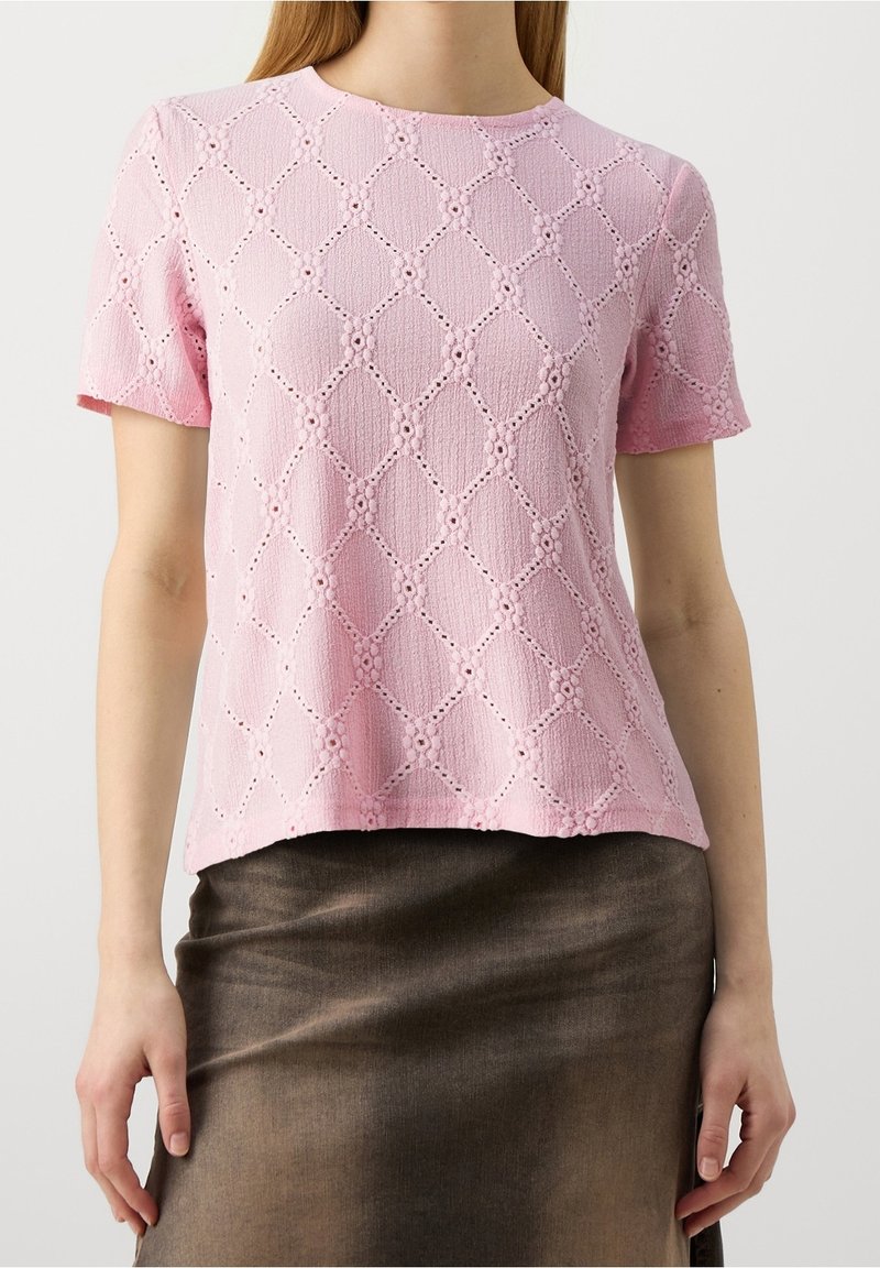 Femme portant un haut texturé rose à manches courtes avec un motif en losange et une jupe marron, debout avec les bras détendus le long du corps.