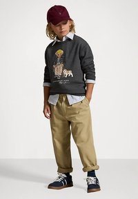 Lapsel on seljas beežid püksid, tumesinised tossud, hall kampsun, millel on karu ja koera muster, triibuline särk ning maroonipunane müts, seistes valge taustaga.