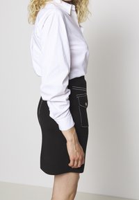 Schwarzer Minirock mit weißen Nähten und Fronttaschendetails, kombiniert mit einem weißen Langarmshirt. Glattes Material mit taillierter Passform.