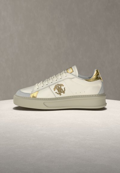 Λευκό αθλητικό παπούτσι χαμηλού ύψους με χρυσές λεπτομέρειες, μπεζ σόλα, κορδόνια και χρυσό λογότυπο στο πλάι, εκτεθειμένο σε φωτεινή επιφάνεια με γκρι φόντο.