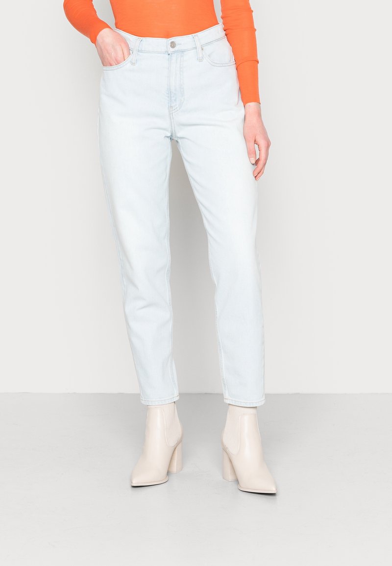Jeans de cintura alta de color azul claro con pierna recta, confeccionados en denim. Llevados con una blusa de manga larga naranja y botas de tobillo de tacón grueso beige.