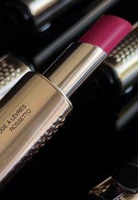 Läppstift i en tub med en polerad silvermetallisk finish, med en djupt rosa kula och texturerade detaljer. Svart bakgrund framhäver produkten.